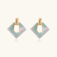Geometric square enamel earrings with marbled turquoise and lavender pattern, gold U-shaped connector on cream background.