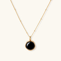Circular black agate pendant on gold-plated sterling silver chain against cream background, showing modern minimalist design.