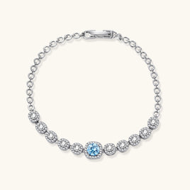 Sterling silver tennis bracelet with round halo-set moissanite stones and blue center stone on cream background.