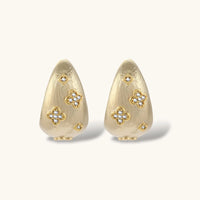 Curved gold-plated hoop earrings with four-leaf clover and diamond-shaped crystal details on white background