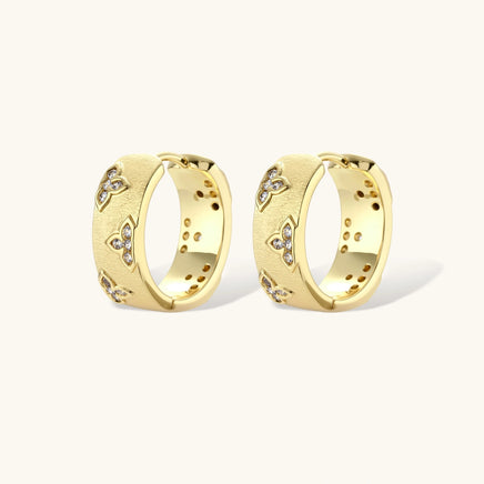 Gold-plated hoop earrings with textured surface and raised floral motifs set with clear zirconia stones on cream background.
