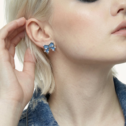 Close-up of blue denim bow stud earring with silver-tone outline and clear zirconia details worn on the ear | Olaty