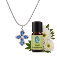 Silver cross diffuser necklace with blue denim detailing displayed beside an amber essential oil bottle with white flowers | Olaty